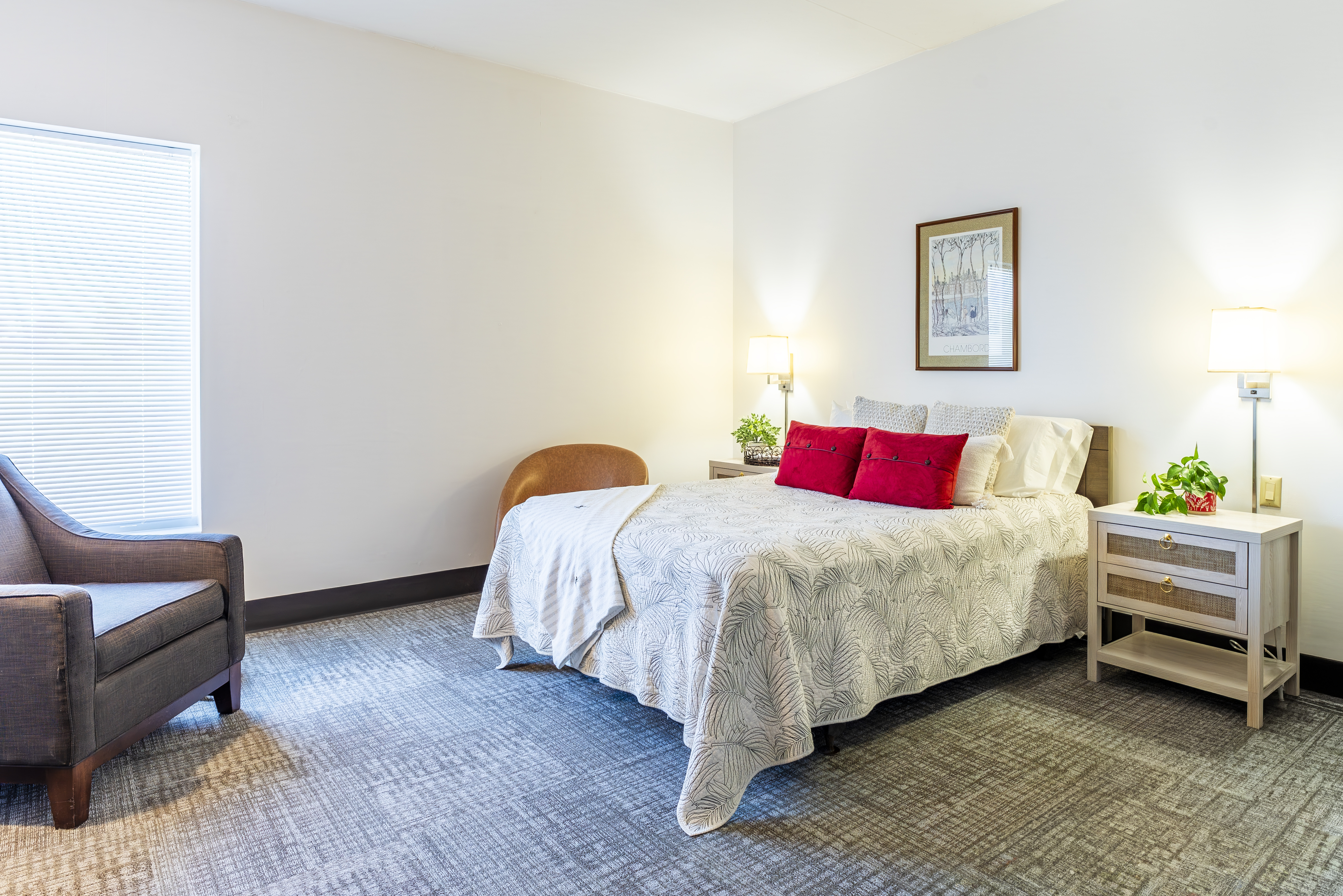 Queen bed with red accent pillows and armchair in furnished room at The Residences, Hadley, MA